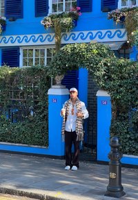 A person in a white shirt and dark trousers stands outside a blue building adorned with decorative planters. They are wearing a patterned scarf and sunglasses.
