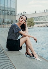 Csíkos rövidujjú felső, fekete szoknya és két tónusú hegyes sarkú cipő (fehér és fekete), amelyet egy víz mellett ülő személy visel.