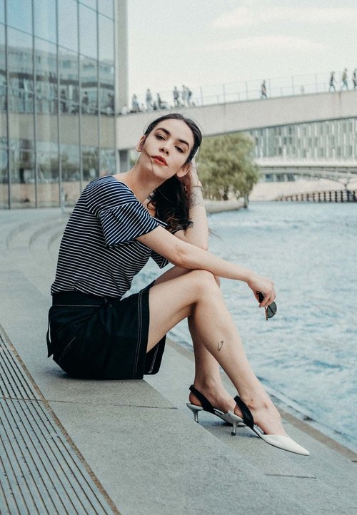 Csíkos rövidujjú felső, fekete szoknya és két tónusú hegyes sarkú cipő (fehér és fekete), amelyet egy víz mellett ülő személy visel.