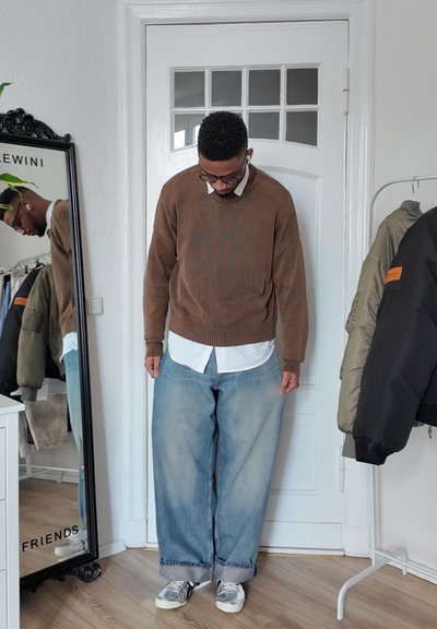 Suéter marrón texturizado sobre una camisa blanca de cuello, combinado con jeans de mezclilla anchos de color azul claro y zapatillas grises. Espejo reflectante en el fondo.