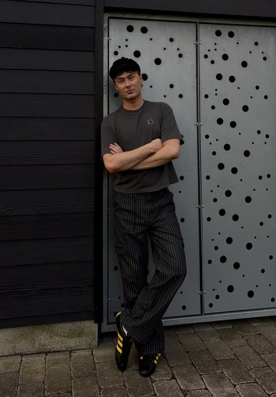 Modelo vistiendo una camisa gris de manga corta y pantalones negros a rayas con un corte relajado, de pie al lado de una puerta gris con recortes circulares.