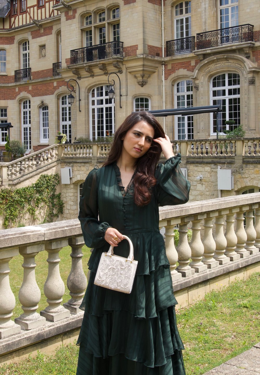 Vestido verde de mangas largas en capas con un tejido texturizado. Bolso blanco estructurado con diseño floral en relieve. Balustrada y edificio histórico de fondo.