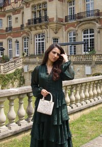 Vestido verde de mangas largas en capas con un tejido texturizado. Bolso blanco estructurado con diseño floral en relieve. Balustrada y edificio histórico de fondo.