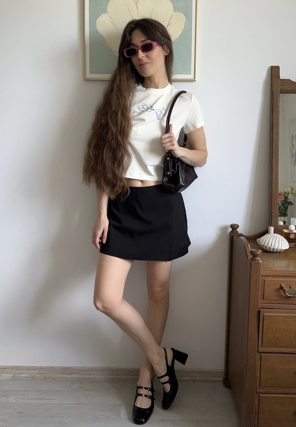 Bijeli skraćeni T-shirt s plavim vezom, crna mini suknja, crne čvrste potpetice i mala tamna torbica. Duga, valovita kosa i ružičaste naočale za sunce.