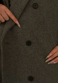 Szürke gyapjúkabát fekete gombokkal és bevágott gallérral. Egy kéz, amelynek hosszú, ápolt körmei vannak, egy gombra mutat.