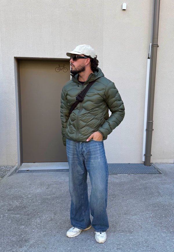 Hombre con chaqueta acolchada verde, vaqueros anchos, zapatillas blancas, gorra blanca y gafas de sol, de pie junto a una pared beige con una puerta marrón.