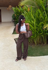 Femme aux longs cheveux tressés et fleur blanche porte un débardeur blanc, un pantalon marron, des sandales noires et tient un sac à main tissé marron près de plantes tropicales.
