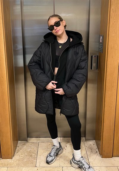 Chaqueta acolchada negra tipo puffer, combinada con un top corto negro y leggings. Los accesorios incluyen gafas de sol negras y zapatillas deportivas grises.