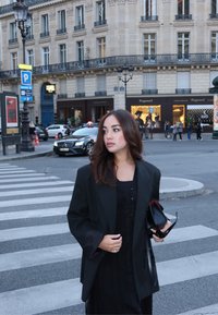 Czarna, oversize marynarka z szerokimi rękawami na czarnej sukience, trzymająca czarna torebkę i okulary przeciwsłoneczne, z miejskim tłem w tle.