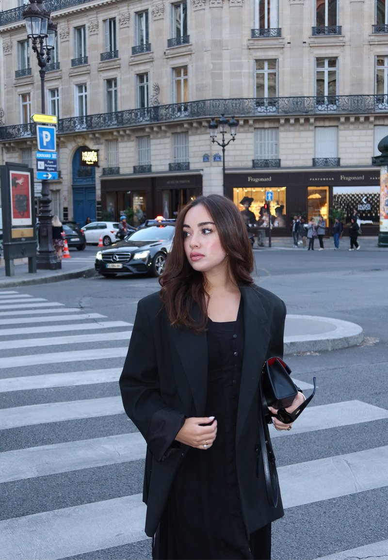 Blazer nero oversize con maniche larghe sopra un vestito nero, che tiene una borsa nera e occhiali da sole, sullo sfondo di una strada di città.
