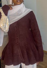 Pull peplum bordeaux avec fermeture à boutons, tissu texturé et ornements de couleur claire. Écharpe blanche drapée sur l'épaule.