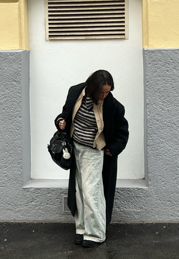 Donna con un lungo cappotto nero, maglione a righe, pantaloni chiari e scarpe nere, che tiene in mano una borsa nera con un portachiavi bianco, in piedi contro un muro grigio.