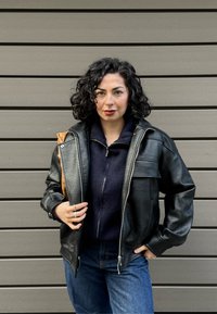 Veste en cuir noir à finition texturée, fermeture éclair et large col ; associée à un haut zippé bleu marine et un jean en denim bleu.