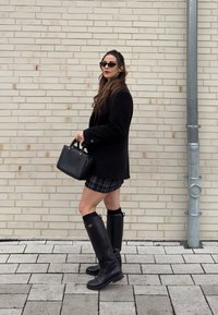 Γυναίκα με μαύρο σακάκι, καρό φούστα, μαύρες μπότες μέχρι το γόνατο, κρατώντας μαύρη τσάντα, στεκόμενη μπροστά σε μπεζ τοίχο από τούβλα με μεταλλικό σωλήνα κοντά.