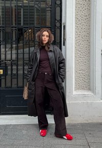 Femme portant un manteau en cuir noir, un pantalon foncé et des baskets rouges, debout les mains dans les poches près d'une grille en métal noir et d'un mur blanc texturé.