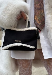 Main tenant un sac à main noir Emily & Noah avec bordure en peau de mouton blanche, portant un manteau en fourrure blanc, une cravate à motifs et des collants en dentelle blanche.