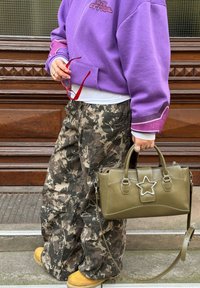 Sweat-shirt violet avec un motif graphique, pantalon camouflage, bottes marron épaisses, et un sac à main vert olive avec un accent en forme d'étoile, porté en bandoulière.
