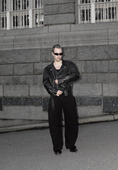 Chaqueta de moto de cuero negro con herrajes plateados, llevada sobre una camiseta negra. Combinada con pantalones negros holgados y gafas de sol reflectantes.