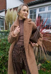 Brown, fluffy coat worn over a beige turtleneck and brown leopard print trousers. Holding a dark brown bag. Featuring green plants and a brick background.