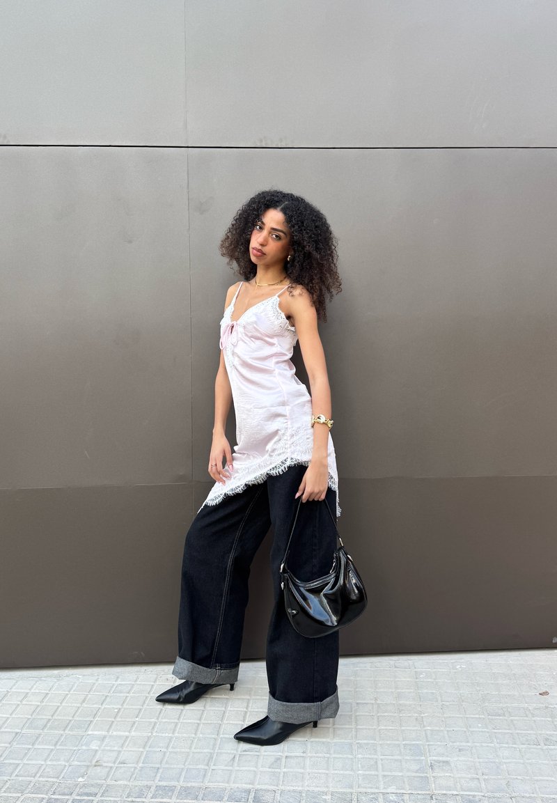 Femme aux cheveux bouclés portant un haut blanc sans manches et un pantalon large noir, tenant un sac à main noir, debout devant un mur gris.