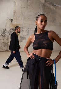 Top cropped de renda preta com decote alto. Saia preta transparente com bolsos e cinto. A modelo segura uma bolsa preta com uma alça azul.