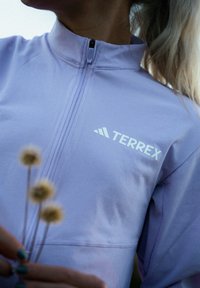 Lys lilla Terrex zip-up jakke med høj krave og logo på fronten. Glat tekstur, tætsiddende design og diskrete stikninger.