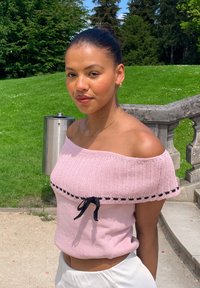 Rosa maglietta a maglia off-the-shoulder con rifiniture nere e dettaglio a fiocco, dalla forma attillata e dalla texture morbida. Abbinata a pantaloni chiari.