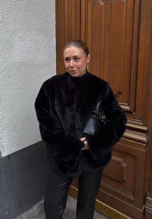 Chaqueta de piel sintética negra con mangas anchas, usada sobre un top negro, combinada con pantalones negros. Pequeño bolso negro con detalles dorados. Fondo de puerta de madera.