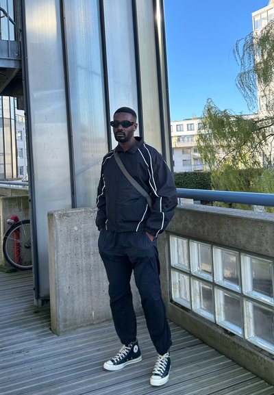 Chaqueta deportiva negra con franjas blancas, pantalones negros a juego y zapatillas altas negras. Fondo de un edificio de cristal, con características de hormigón.