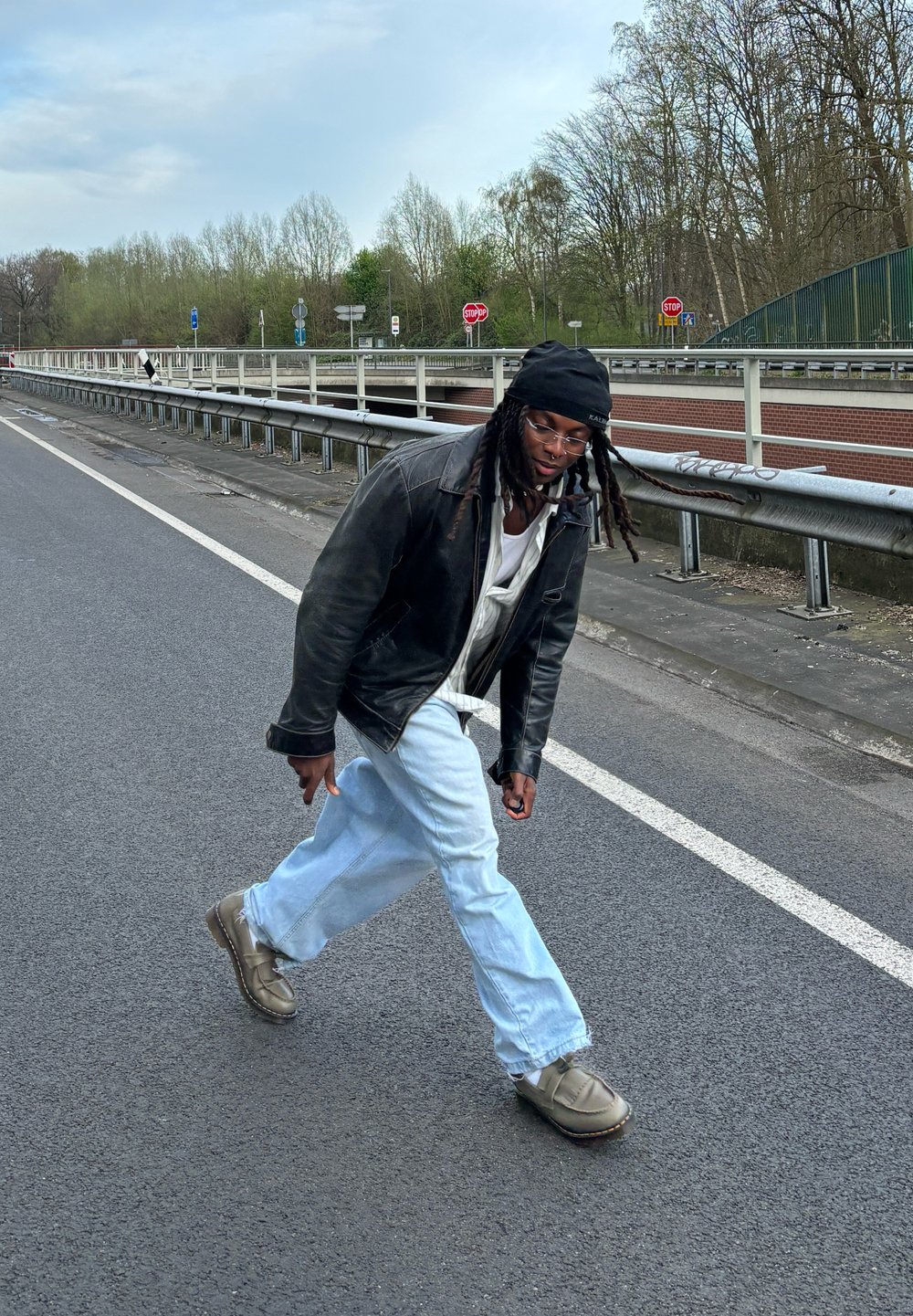 Crna kožna jakna, bijela košulja, svijetloplave široke traperice i smeđe cipele na slip-on. Hoda po cesti s zelenilom i prometnim znakovima u pozadini.