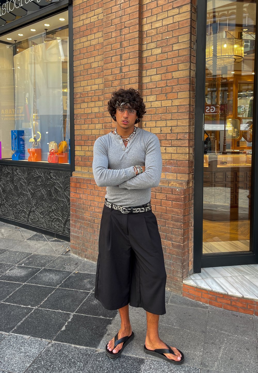 Grå langermet skjorte, svarte vide shorts og svarte flip-flops. Tilbehør inkluderer flere armbånd og et dekorativt belte. Murvegg som bakgrunn.