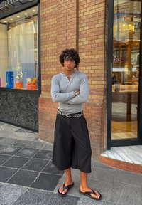 Grå langermet skjorte, svarte vide shorts og svarte flip-flops. Tilbehør inkluderer flere armbånd og et dekorativt belte. Murvegg som bakgrunn.