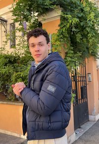 Giubbotto imbottito blu navy con design trapuntato, chiusura con zip e patch "POLO CLUB" sulla manica sinistra, abbinato a una camicia azzurra sottostante.