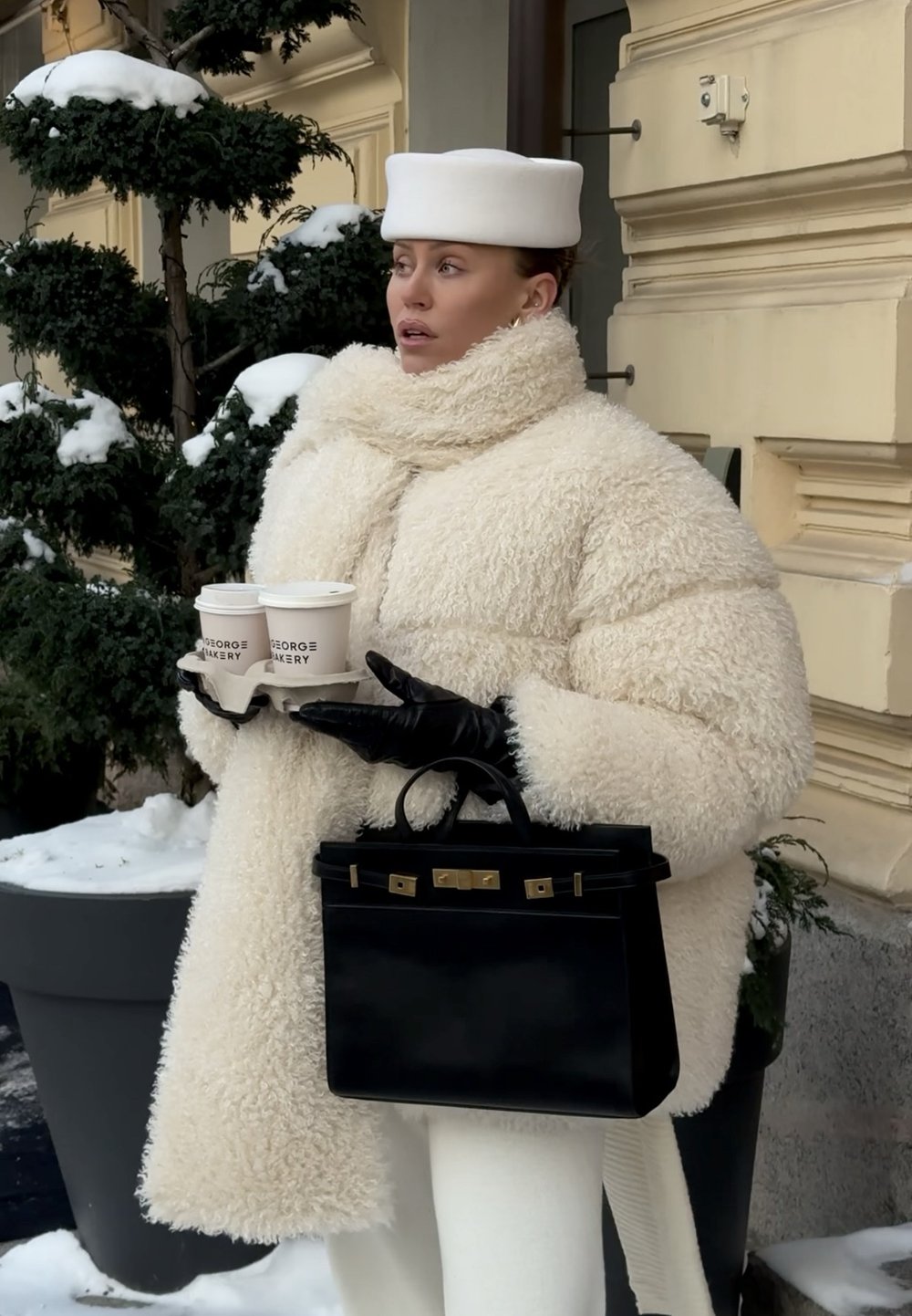Personne en manteau de fourrure blanc et chapeau tient deux gobelets de café à emporter et un sac à main noir, debout à l'extérieur près de plantes couvertes de neige.