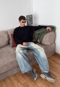 Svart oversize tröja, blekta vida blå jeans, gråa klumpiga sneakers, sittande på en strukturerad beige soffa med en grön pläd över.