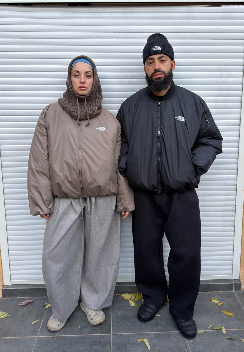 Deux personnes se tiennent devant un volet blanc texturé. L'une porte une doudoune marron et un pantalon large gris, l'autre une doudoune noire et un pantalon noir.