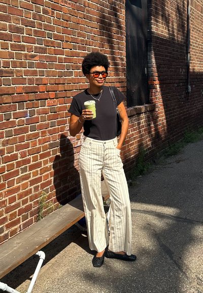 Camisa negra de manga corta, pantalones anchos a rayas en color crema, zapatos negros. Sosteniendo una bebida verde, de pie contra una pared de ladrillo.