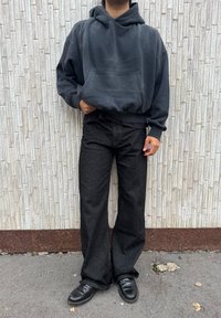 Personne portant un sweat à capuche sombre et surdimensionné, un jean noir à jambes larges et des chaussures en cuir noires, debout contre un mur en briques texturé.