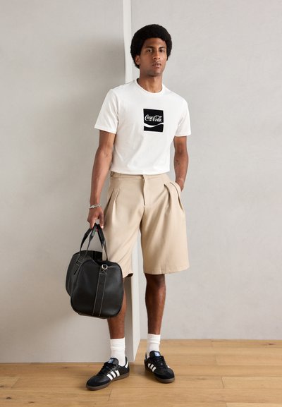 Camiseta blanca con gráfico de Coca-Cola, pantalones cortos de lino beige con pliegues, bolso de cuero negro, zapatillas negras con rayas blancas y calcetines blancos.