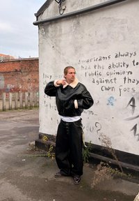 Personne portant une veste en cuir noire et un pantalon ample se tient près d'un mur blanc avec un graffiti délavé dans un environnement urbain extérieur.