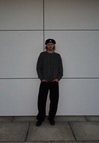 Grau gestrickter Pullover, schwarze weit geschnittene Hose und ein schwarzes Barett mit weißem Logo. Große, schlichte Wand im Hintergrund, minimaler Kontrast.