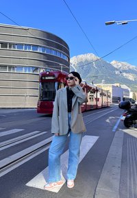 Personne portant des lunettes de soleil, un blazer oversized et un jean se tient sur un passage piéton devant un tram rouge avec des montagnes et un bâtiment moderne en arrière-plan.