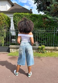 Jeans culotte in denim blu chiaro con orlo grezzo, indossati con un top bianco a spalle scoperte e sneakers bianche con dettagli verdi. Sfondo recintato.