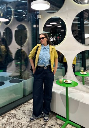 Una persona con una chaqueta amarilla y una camiseta azul claro está de pie frente a espejos circulares, lleva gafas de sol negras, jeans azules anchos y zapatillas.