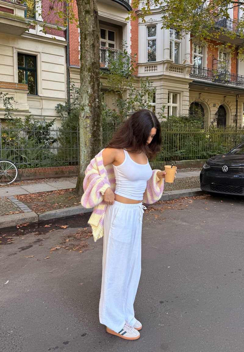 White wide-leg trousers, a fitted white tank top, and a pastel striped cardigan in pink and yellow. Holding a coffee cup outside.