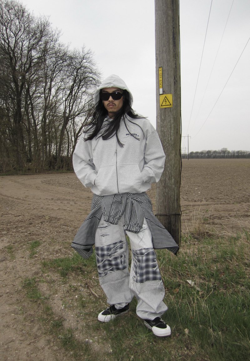 Personne portant un sweat à capuche gris, un pantalon patchwork déchiré et des lunettes de soleil, debout près d'un poteau électrique dans un champ rural sous un ciel couvert.