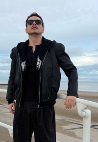 Crna kožna bomber jakna s kapuljačom, s bijelim grafičkim detaljima. Nošena preko crnog henley topa i širokih kariranih hlača.