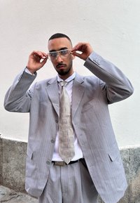 Ανοιχτό γκρι κοστούμι με ρίγες και μεγάλα κουμπιά, με ένα σχέδιο γραβάτα και καθαρά γυαλιά. Τοποθετημένο σε απλό λευκό φόντο.