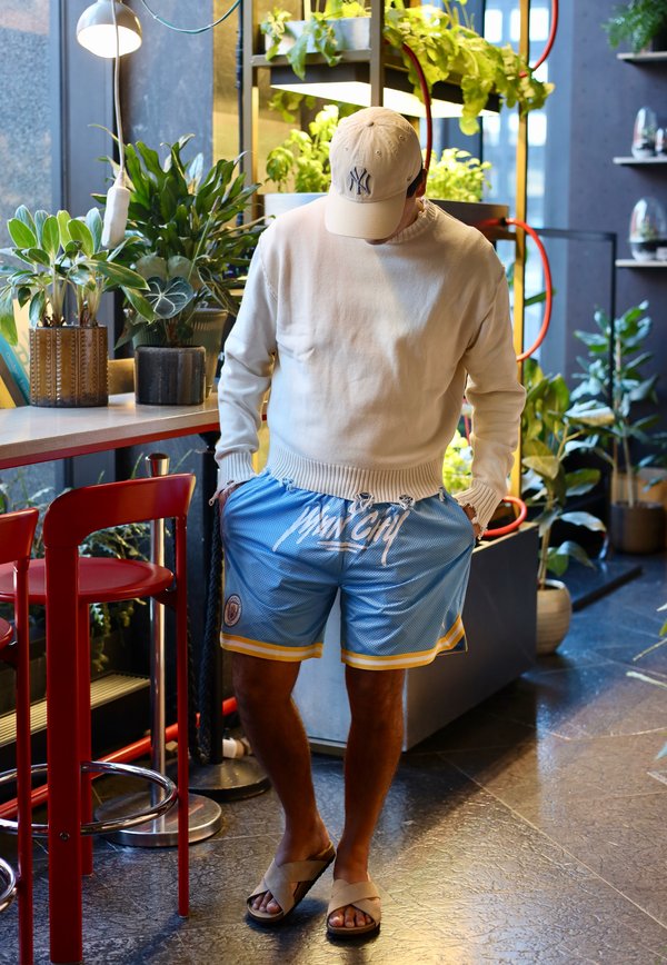 Persoană îmbrăcată într-un pulover bej, pantaloni scurți albaștri „Man City”, șapcă bej și sandale, stând în interior lângă scaune roșii și plante verzi diverse.