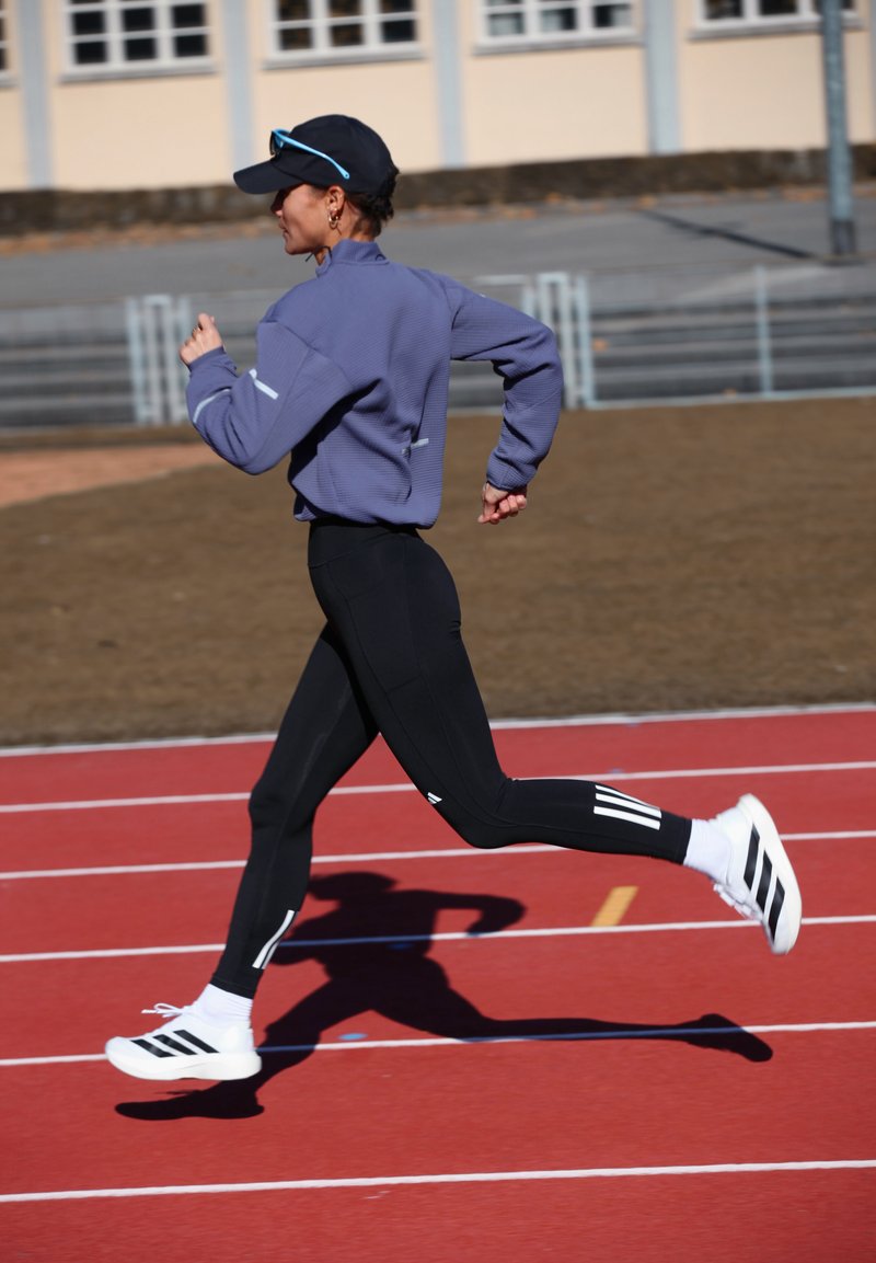 Woman in black leggings and white sneakers running on a red track during daytime, wearing a purple jacket and black cap with sunglasses.